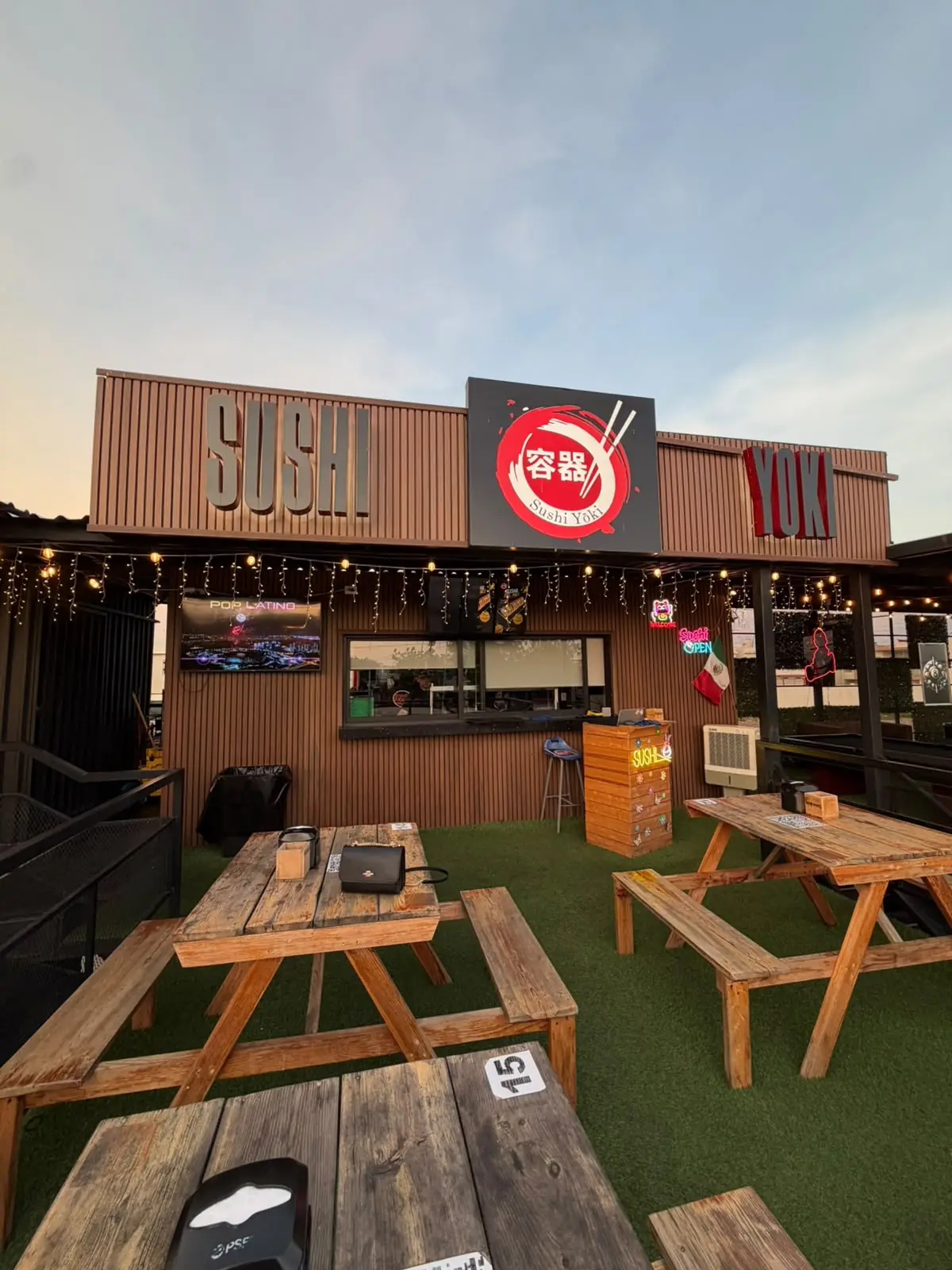 Contenedor de Sushi Yōki con su letrero rojo y mesas de madera sobre pasto